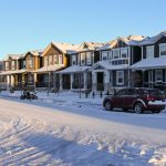 Suburban homes and street covered in snow after a blizzard. The sky is clear now, and the sun is rising on these medium-sized homes of various building styles. There are cars and a motorbike parked in the street.