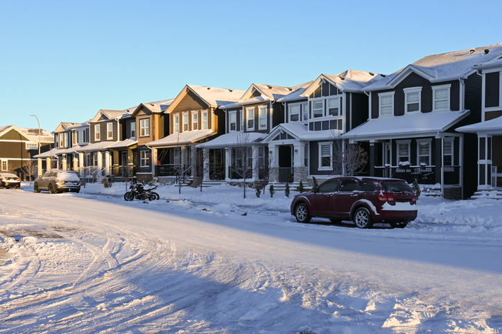 Suburban homes and street covered in snow after a blizzard. The sky is clear now, and the sun is rising on these medium-sized homes of various building styles. There are cars and a motorbike parked in the street.