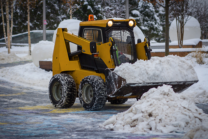 A big orange excavator shovels snow from the road and clears the sidewalk. A snowplow clears roads in the city from snow in winter. Cleaning after snowfalls and blizzards.